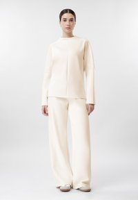 Mujer de pie, vestida con una blusa de manga larga color marfil y pantalones de pierna ancha con paneles de encaje, combinados con zapatos beige sobre un fondo blanco liso.