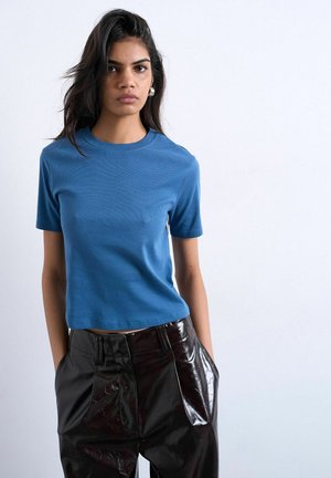 Woman with long dark hair wearing blue short-sleeve shirt and shiny black pants, standing with hands in pockets against white background.