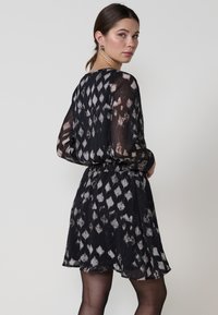 Schwarz-weiß gemustertes Kleid mit langen, transparenten Ärmeln, tailliertem Schnitt, ausgestelltem Rock und Rundhalsausschnitt. Der transparente Stoff betont die Bewegungen.