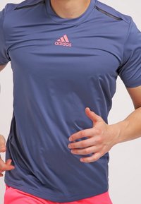 Camiseta deportiva azul de material ligero con mangas cortas, con detalles en negro en los hombros y un logo rosa de Adidas en el pecho.