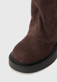 Braune Wildleder-Stiefelette mit einer abgerundeten Zehenkappe und einer schwarzen Plateau-Sohle. Die Textur ist weich und zeigt eine Naht entlang der Nähte.