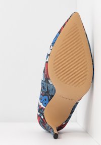 Chaussure à talon haut pointue avec imprimé style bande dessinée en rouge, bleu, blanc et noir, vue de la semelle avec semelle extérieure texturée couleur fauve.