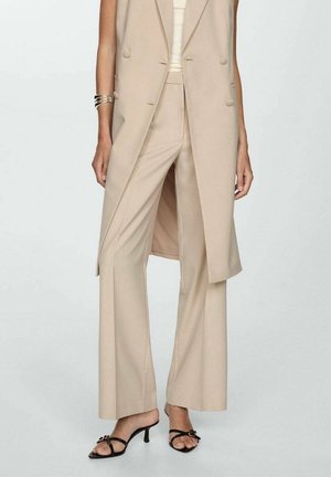 Beige maßgeschneiderte, weit geschnittene Hose mit passendem ärmellosem langem Blazer, schwarze Riemchen-Absatzsandalen und silbernes Manschettenarmband am linken Handgelenk.