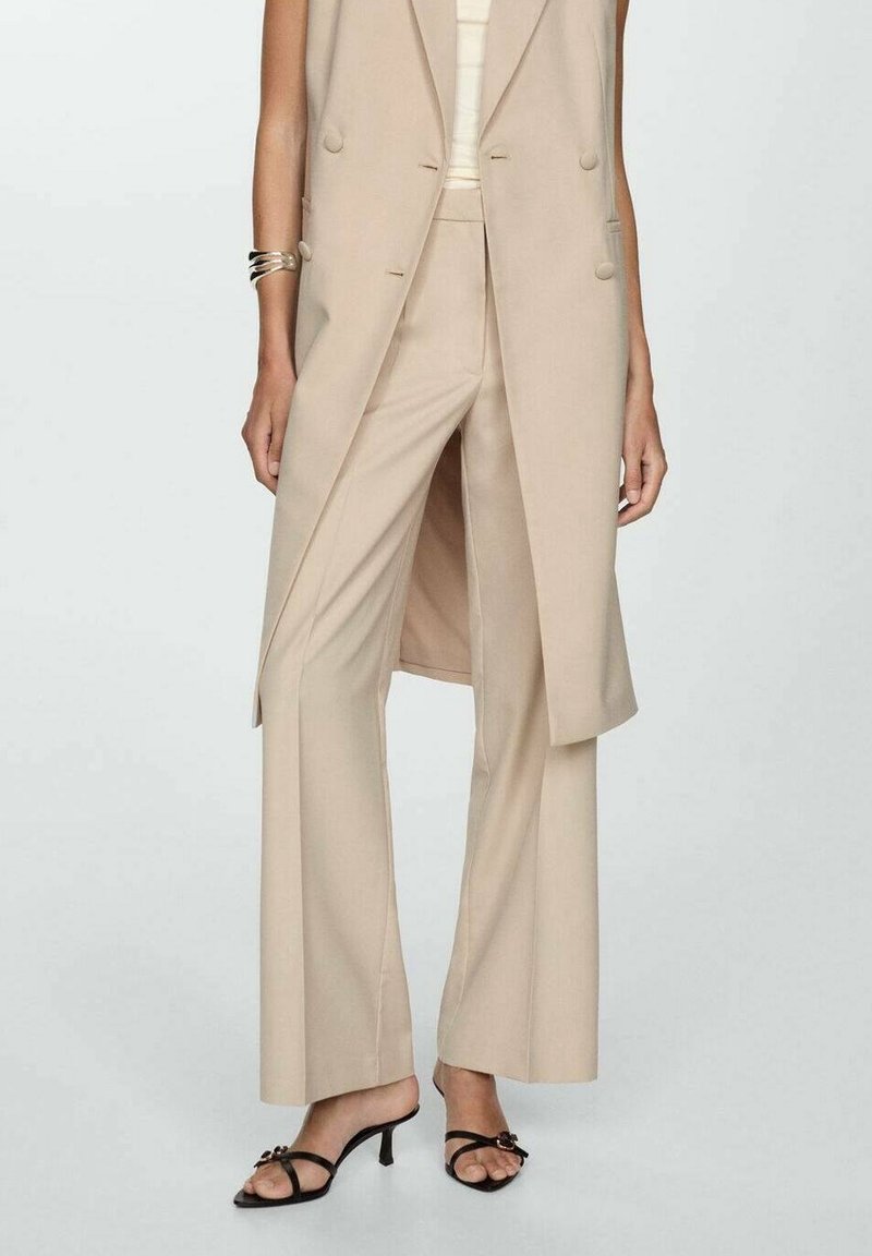 Pantalon beige à jambes larges ajusté avec blazer long sans manches assorti, sandales noires à talons avec lanières et bracelet manchette argenté au poignet gauche.