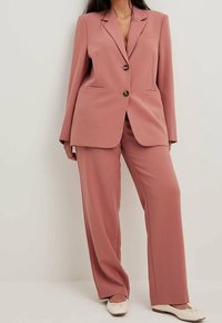 Rosa skräddarsydd kavaj med två framfickor och två knappar, i kombination med matchande vida byxor. Strukturerad tyg, minimal detaljer.