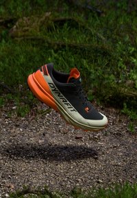 Ελαφρύ αθλητικό παπούτσι τρεξίματος με μαύρο επάνω μέρος, πορτοκαλί λεπτομέρειες και υφασμάτινη γκρι σόλα. Ο σχεδιασμός τονίζει την απόδοση και τη σταθερότητα.