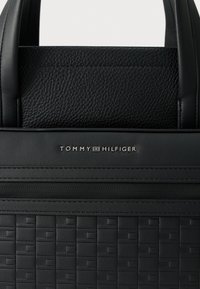 Fekete bőrből készült kézitáska, mely texturált, szövött mintával, sima díszítésekkel és dombornyomott márkajelzéssel rendelkezik. Két fogantyúval és cipzáras záródással ellátva.