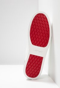 White sneaker sole with textured red rubber pattern. Smooth edges and subtle branding on the white side, angled against a light background.