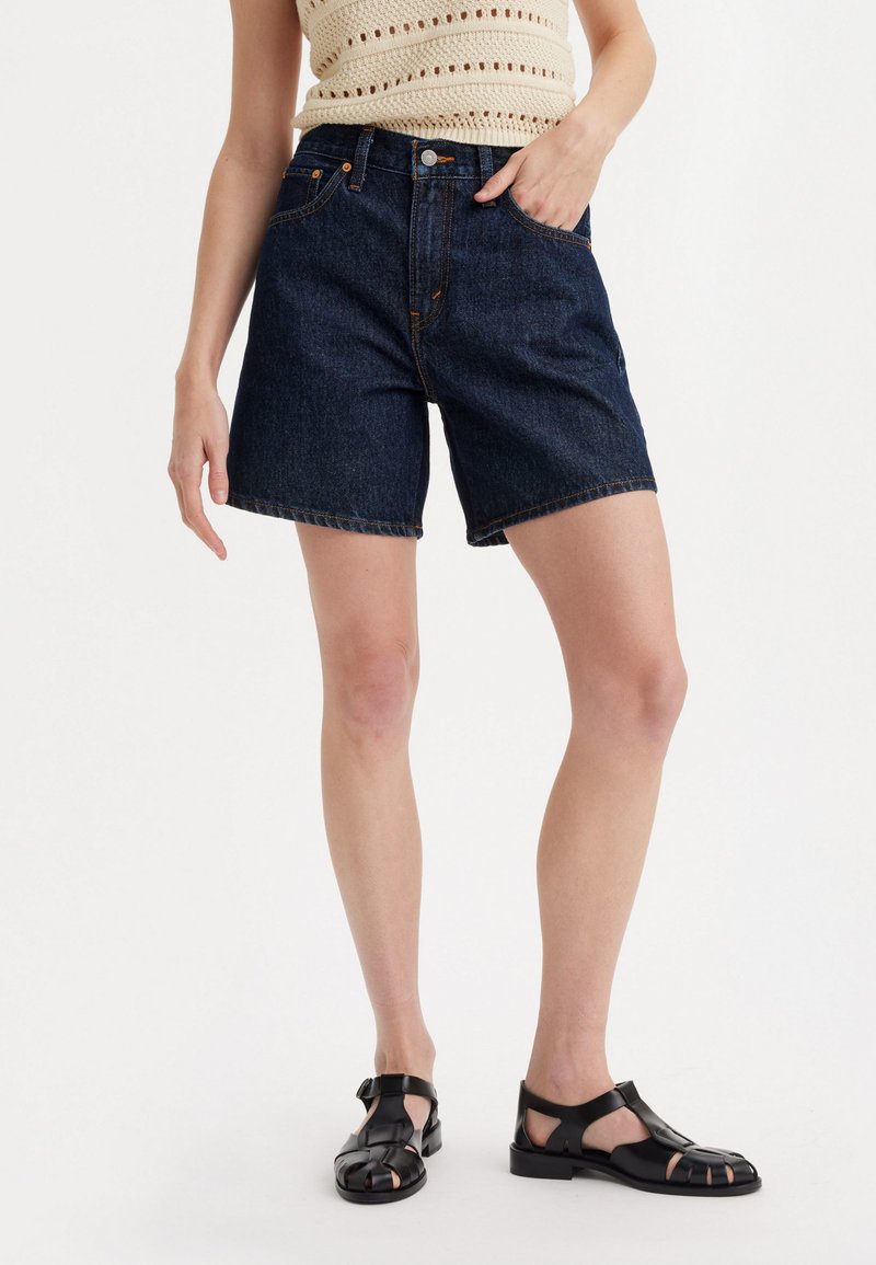 Pantalones cortos de mezclilla en azul oscuro con cintura alta, con costuras en contraste, bolsillos laterales y un corte recto. Llevados con sandalias negras.