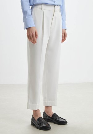 Pantaloni bianchi su misura con pieghe, caratterizzati da una vestibilità rilassata e orli, abbinati a mocassini neri in vernice. Il tessuto appare liscio e strutturato.