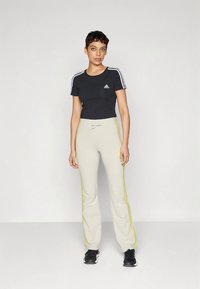 Γυναίκα standing wearing μαύρο Adidas t-shirt, μπεζ Adidas track pants με κίτρινες ρίγες και μαύρα sneakers, μπροστά σε λευκό φόντο.