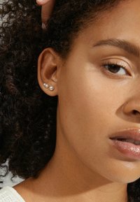 Trois petites boucles d'oreilles en perles dans un design de taille graduée, mises en valeur par des cheveux bouclés et une peau lisse. La lumière naturelle accentue la texture.