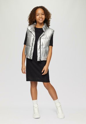Silberner, gepolsterter Westen über einem schwarzen Kleid, mit einem Reißverschluss vorne und hohem Kragen. Das Model trägt weiße Sneakers im High-Top-Stil und Socken.