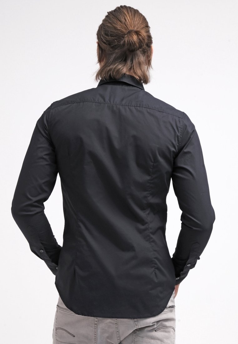 Homme avec cheveux attachés portant une chemise noire ajustée à manches longues et un pantalon de couleur claire, vu de dos sur un fond blanc.