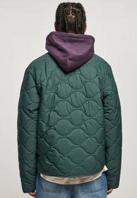 Chaqueta de plumas verde acolchada con un patrón texturizado, acompañada de una sudadera con capucha morada debajo, vista desde la parte de atrás.