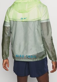 Letvægts Nike Trail-jakke i neongrøn med grå og mørkegrønne accenter. Har hætte, teksturerede mønstre samt blå logo og tekst.