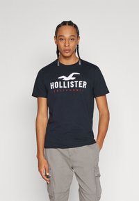 Melna kokvilnas T-krekls ar uzrakstu "HOLLISTER CALIFORNIA" baltā un sarkanā krāsā, ar apaļu kakla izgriezumu, īsām piedurknēm un brīvu piegriezumu.