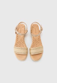 Des sandales beiges avec une lanière en jute tressé et une lanière de cheville en cuir lisse. La semelle intérieure texturée présente un motif de feuilles. Accents en matériel bronze.
