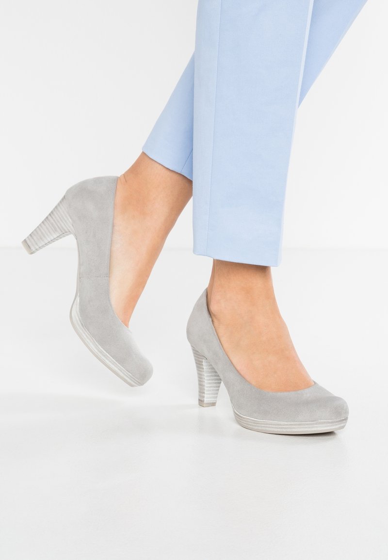 Femme portant un pantalon bleu clair et des chaussures à talons hauts en suède gris à talons empilés, debout sur un sol blanc devant un fond blanc.