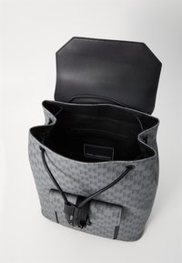 Sac fourre-tout en textile gris avec un rabat en cuir noir et une fermeture à cordon. Il dispose d'une étiquette de logo à l'intérieur et d'une poche avant avec un fermoir en métal.