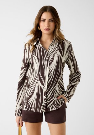 Kobieta w brązowo-białej koszuli na guziki z abstrakcyjnym wzorem oraz brązowych szortach, trzymająca beżową torebkę na białym tle.