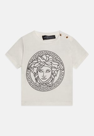 T-shirt en coton crème avec un graphique de Méduse noir et un motif de clé grecque circulaire. Comprend des boutons dorés sur une épaule.