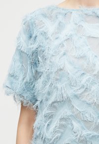 Ανοιχτό γαλάζιο διαφανές ύφασμα με υφή και φτερωτές λεπτομέρειες στο τελείωμα, που δημιουργούν ένα στρωματοποιημένο, απαλό και λεπτεπίλεπτο σχέδιο σε ένδυμα με κοντά μανίκια.