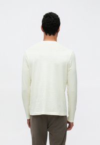 Langärmeliges T-Shirt in Off-White mit rundem Ausschnitt und strukturierter Stoffqualität. Kombiniert mit brauner Hose, die eine entspannte Passform zeigt.