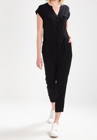 Combinaison courte noire à manches courtes avec bouton sur le devant, deux poches poitrine, taille cintrée et jambes fuselées, associée à des baskets blanches.