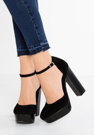 Zapatos de tacón plataforma de ante negro con correa en el tobillo y tacón cuadrado. Los zapatos tienen una textura suave y diseño de punta redondeada.