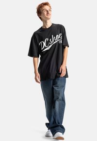 Černé oversized tričko s bílým logem "DC shoe co.", nošené s tmavě modrými volnými džíny a bílými teniskami, na světlém pozadí.