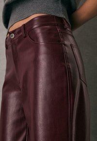 Burgundy læderbukser med høj talje, der har en glat tekstur, for-lommer og en enkelt knaplukning med subtile syninger.