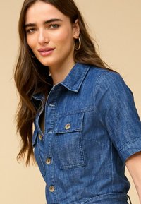 Vestito camicia in denim blu con maniche corte, chiusura a bottoni frontale, tasche sul petto e colletto strutturato. Presenta un tessuto testurizzato.