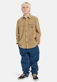 Šviesiaplaukis vaikas stovi su rankomis kišenėse, vilkintis šviesiai rudą mygtukinį kordūro marškinių, laisvas mėlyną džinsus ir juodus sportinius batelius ant balto fono.