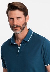 Polo tričko v tmavě tyrkysové barvě s texturovanou tkaninou, s pevným límcem a bílými opaskovými akcenty na límci a rukávech.