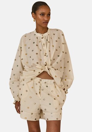 Kobieta ubrana w beżową koszulę na długi rękaw zapinaną na guziki oraz pasujące szorty z wielokolorowymi okrągłymi wzorami, koszula zawiązana w talii, ręce w kieszeniach.
