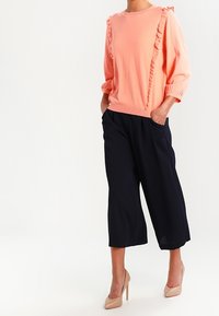 Korálový pulovr s volánkovým detailem podél předních švů, doplněný volnými námořnicky modrými kulotami; modelka má na sobě tělové lodičky.