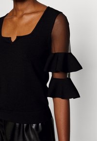 Schwarzes Strickoberteil mit V-Ausschnitt, transparenten Netzärmeln und ausgestellten Rüschendetails an den Ellbogen. Weiche Textur, figurbetontes Design.