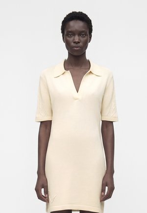 Frau mit kurzen lockigen Haaren, die ein kurzärmeliges cremefarbenes Kleid mit Kragen und V-Ausschnitt trägt, steht vor einem schlichten Hintergrund.