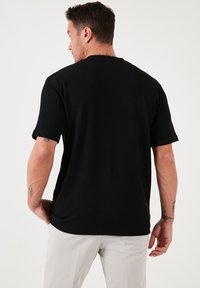 Schwarzes geripptes T-Shirt mit kurzen Ärmeln, lockerer Passform, Rundhalsausschnitt und strukturierter Stoff. Sichtbare Tattoos am Arm des Trägers.