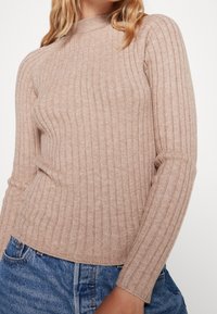 Suéter beige de punto acanalado con cuello alto y mangas largas, con una silueta ajustada y textura suave. Combinado con jeans de mezclilla azul.