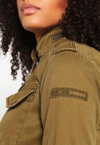 Olivgrüne Jacke mit glatter Textur, ausgestattet mit Schulterpolstern und einem Patch mit der Aufschrift "AKF-1892." Sie hat aufgesetzte Taschen und einen Reißverschluss.