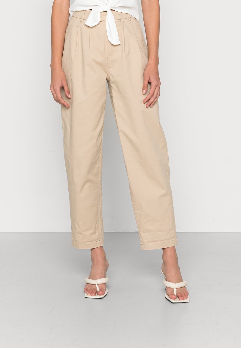 Beige high-waisted trousers with a relaxed fit, featuring front pleats and a tied waistband. Pair of white square-toed sandals completes the look.