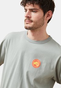 Camiseta de algodón gris con un logo gráfico colorido y redondo que presenta un mapa estilizado y una cruz, cuello redondo y corte relajado.