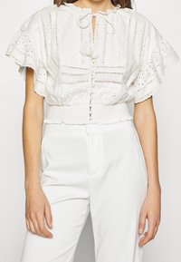 Weiße Bluse mit kurzen, gerafften Ärmeln, die mit Augenstickerei verziert ist und einen Krawattenkragen hat. Knopfleiste vorne und elastischer Bund für eine gute Passform.