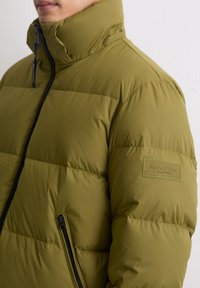 Kurtka puchowa w kolorze oliwkowej zieleni z wysokim kołnierzem, zamkiem błyskawicznym i poziomym przeszyciem; posiada naszywkę z logo na lewym ramieniu.