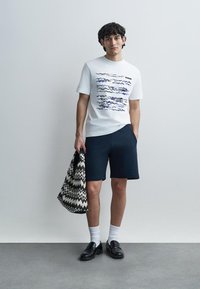 Λευκό βαμβακερό T-shirt με μπλε σχέδιο κυμάτων, συνδυασμένο με σκούρο μπλε σορτς. Κρατάει μαύρη και λευκή τσάντα με ζιγκ ζαγκ. Μαύρα παπούτσια και λευκές κάλτσες.