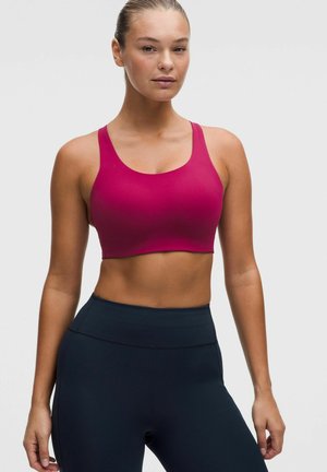 Frau trägt einen leuchtend magentafarbenen Sport-BH und hoch taillierte schwarze Leggings, steht vor einem schlichten hellen Hintergrund.