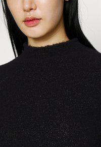 Schwarzer Pullover mit Struktur und hohem Ausschnitt, der einen subtilen Glanz aufweist. Das Material wirkt weich und leicht flauschig, mit einem cropped Saum.