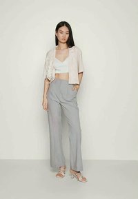 Mulher vestindo uma jaqueta cropped bege, um top cropped branco, calças largas cinzas e saltos brancos com tiras, posando contra um fundo claro e uniforme.